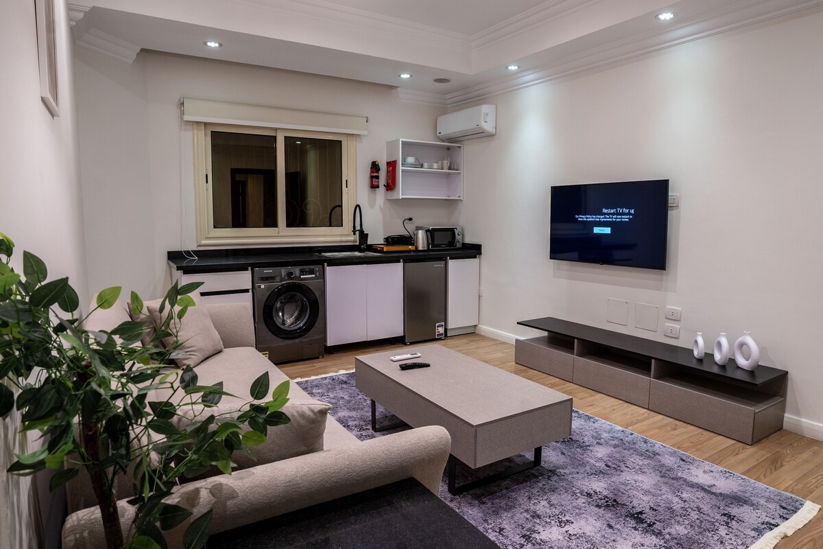 A modern living area features a comfortable sofa positioned across from a wall-mounted smart TV. A sleek coffee table is centered on a soft rug, while a kitchenette with essential appliances is visible in the background. Natural light enters through a large window.