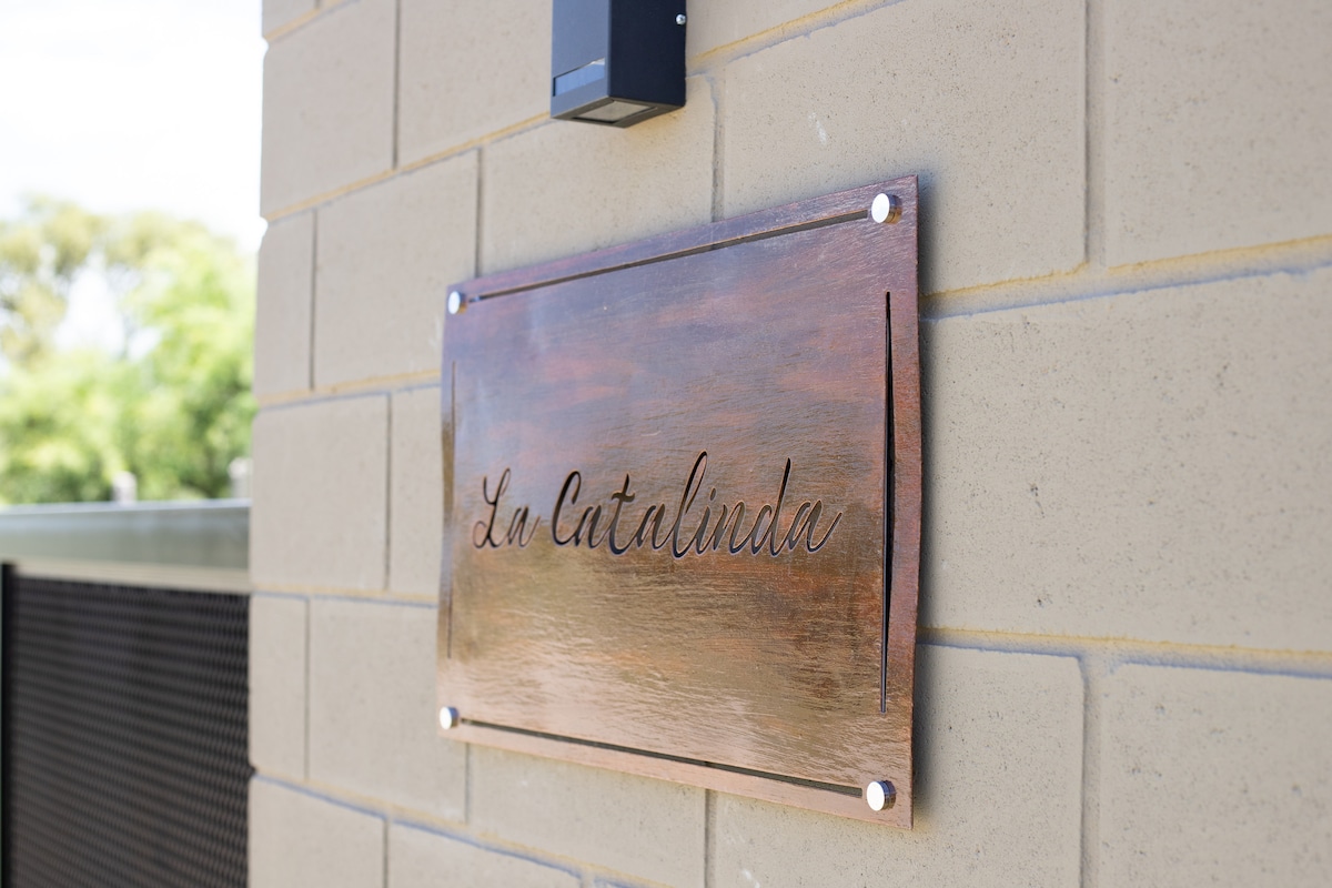 A decorative sign displaying the name 'La Catalinida' is mounted on a smooth beige brick wall. The sign features a polished metal finish, with the text presented in an elegant script, providing an inviting touch to the entryway.