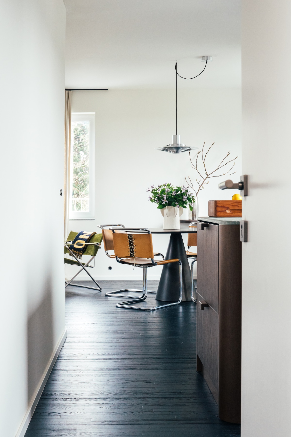 A bright hallway leads to a dining area with a sleek table surrounded by modern chairs. A pendant light hangs above, illuminating a vase of flowers on the table. Natural light filters in through a nearby window, enhancing the open feel of the space.
