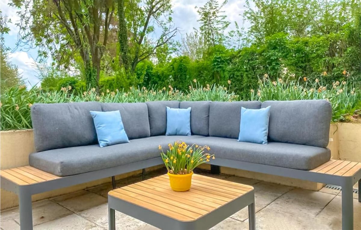 A modern outdoor seating area is featured, comprised of a gray sectional sofa adorned with light blue cushions. A wooden table is positioned in the center, accented by a yellow pot containing vibrant flowers. Lush greenery and trees form a natural backdrop.