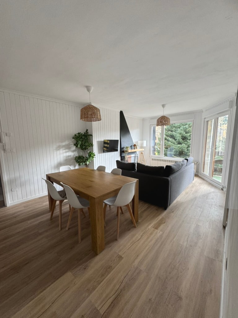 A dining area features a wooden table surrounded by six white chairs. A dark sofa is positioned nearby, with a tall black fireplace adding a modern touch. Natural light enters through large windows, illuminating the wooden floor and creating a welcoming atmosphere.