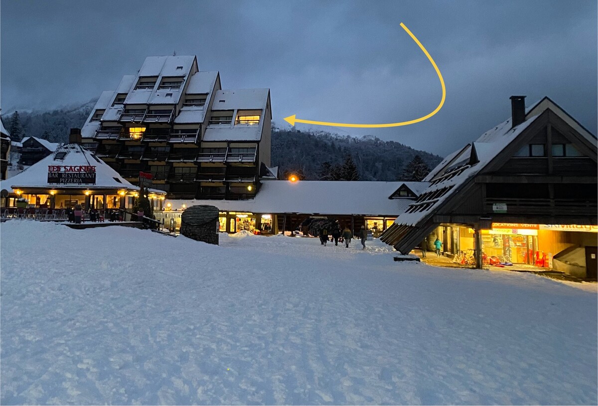 A snow-covered landscape features a modern, multi-level building, with its distinctive sloped roofs visible against a cloudy sky. Nearby, a cozy structure houses shops and restaurants, while a gently illuminated warm glow emanates from the windows, inviting visitors to explore.
