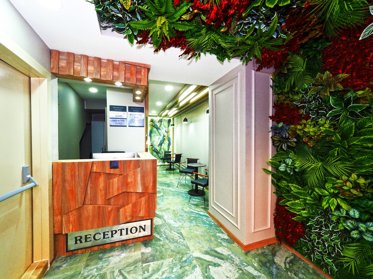 The reception area features a wooden front desk with the word 'RECEPTION' prominently displayed. A vibrant wall of greenery and colorful foliage provides a striking contrast against the polished stone flooring. Small tables and chairs are visible in the background, creating a welcoming atmosphere.