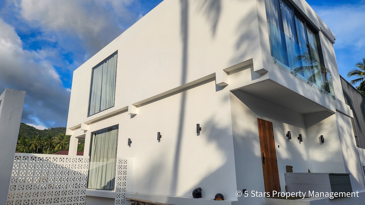 The exterior of a modern two-story villa is showcased, featuring a sleek white facade and large glass windows. The entrance stands welcoming with a well-defined door, and palm trees are visible in the background, providing a sense of the surrounding lush environment.