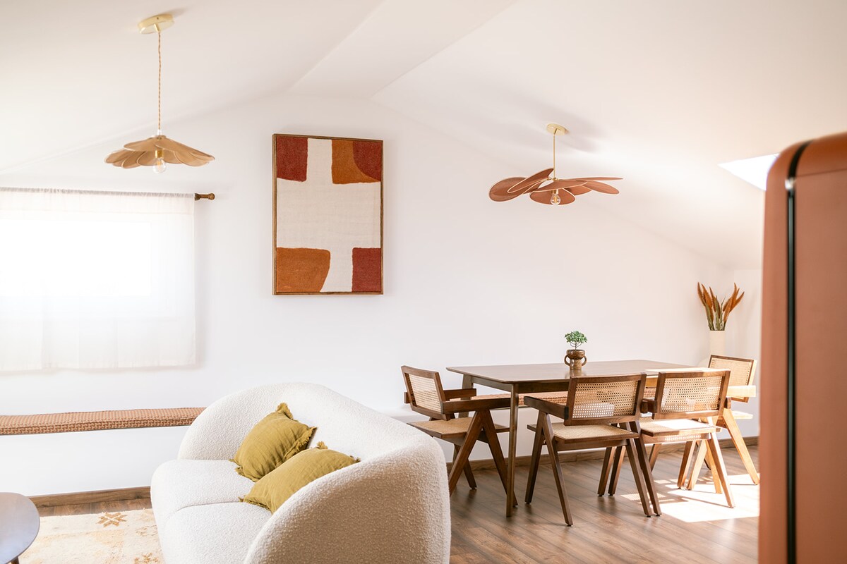 A bright living area features a light-colored sofa with decorative cushions alongside a wooden dining table surrounded by several chairs. Two ceiling fans are present, and natural light filters through a window adorned with light drapes, while a piece of wall art adds a touch of color.