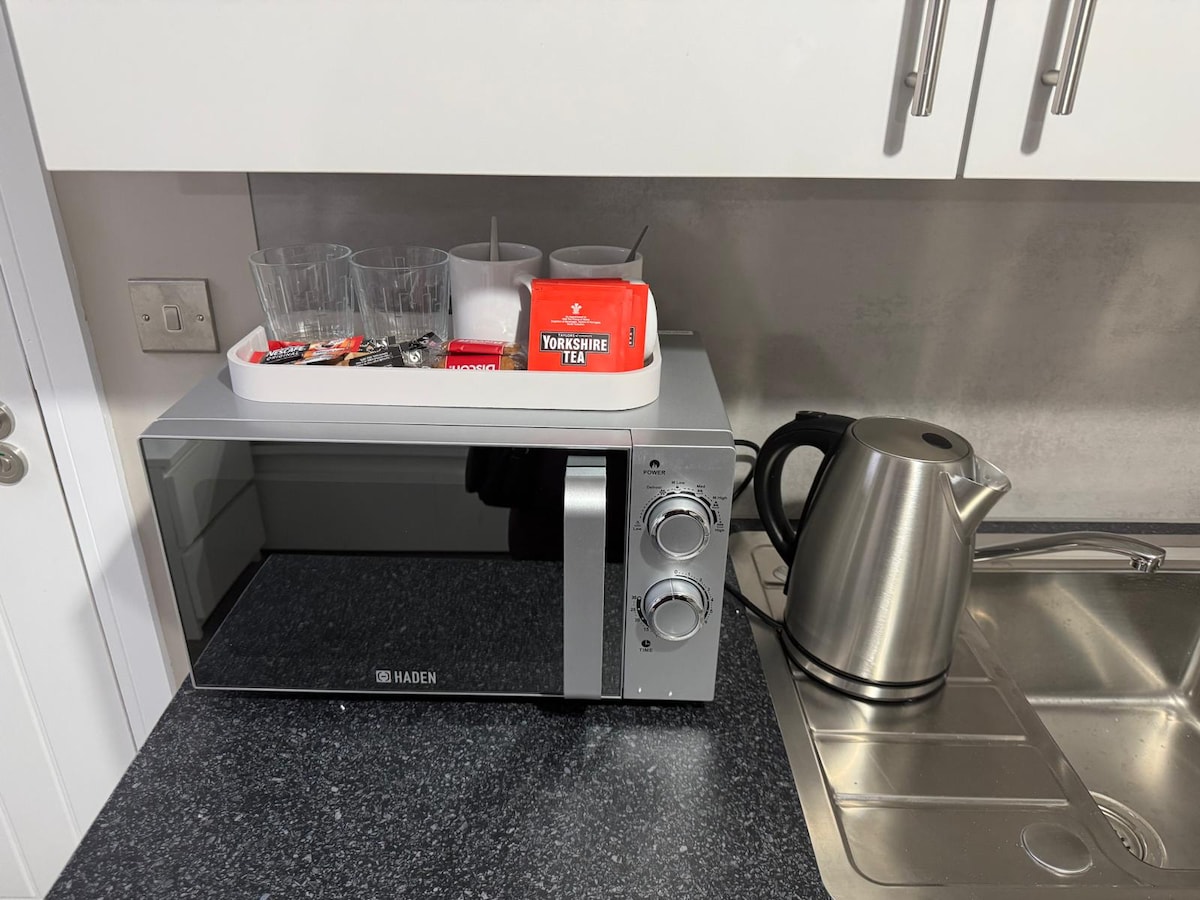 A modern kitchenette corner is equipped with a microwave positioned beside a stainless steel kettle. A white tray holds several glasses and a box of tea. The countertop features a sleek sink set into the dark surface.