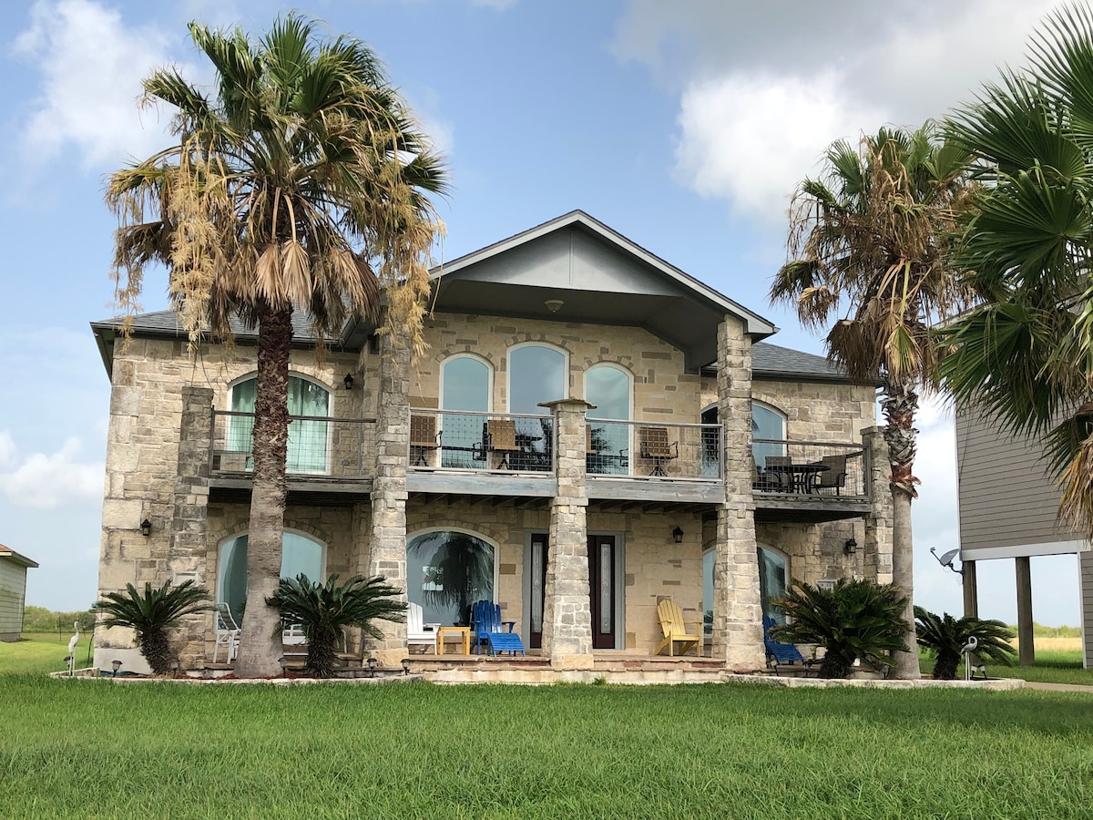 The two-story house is constructed of Texas Hill-Country Limestone, featuring wide arched windows and a balcony with chairs. Palm trees are positioned around the exterior, and the lush green lawn offers a fresh and open atmosphere. The imposing stone structure exudes a sense of spaciousness.