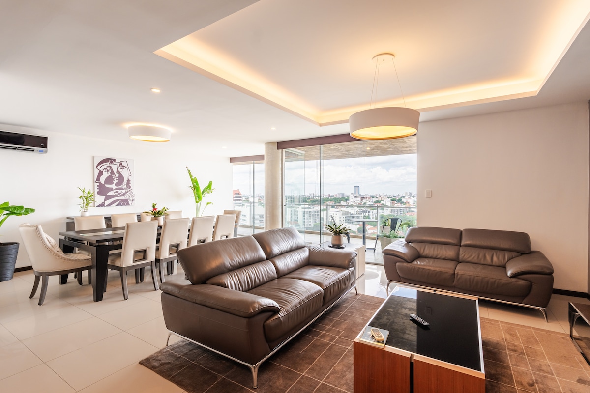 A spacious living area is fitted with two modern leather sofas facing a chic coffee table. Natural light pours through expansive glass windows, revealing a city view. A dining table for six is visible in the background, surrounded by potted plants.