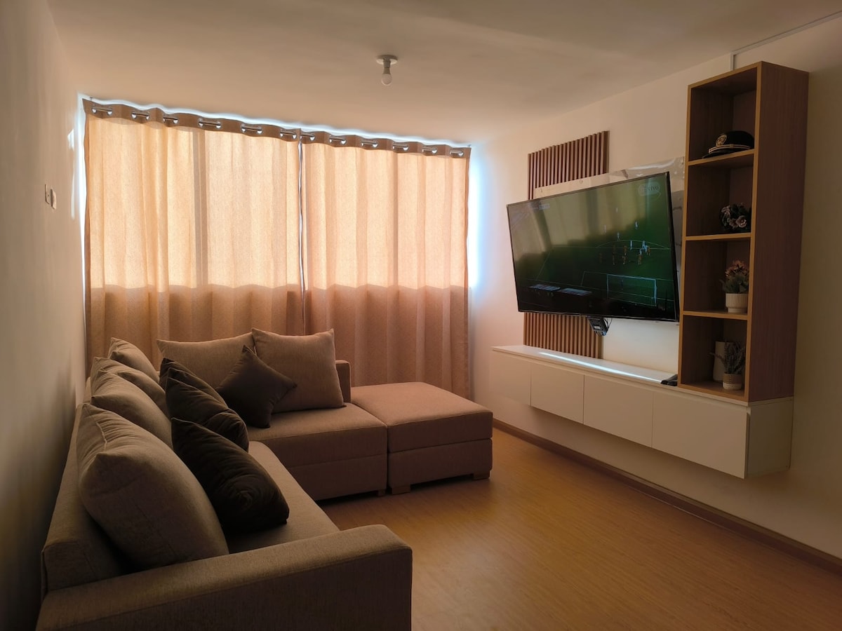 A cozy living area is highlighted by a sectional sofa and a wall-mounted television. Natural light filters through light curtains, creating a warm ambiance. A minimalist entertainment unit is positioned below the TV, complementing the simple design of the space.