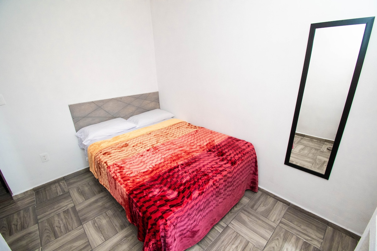 A cozy sleeping area is featured, with a colorful blanket draped over the spacious bed. A large mirror leans against the wall, reflecting the room's light and bright walls. The floor is covered with square tiles, creating a clean and modern aesthetic.
