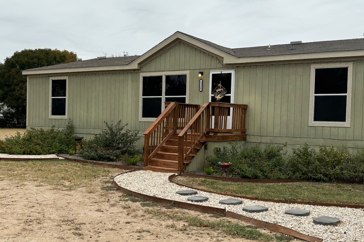 A single-story rental home is presented, featuring light green siding and multiple large windows. A wooden staircase leads to the front door, framed by a landscaped entry with stepping stones and soft greenery. The inviting exterior is complemented by a peaceful surrounding.