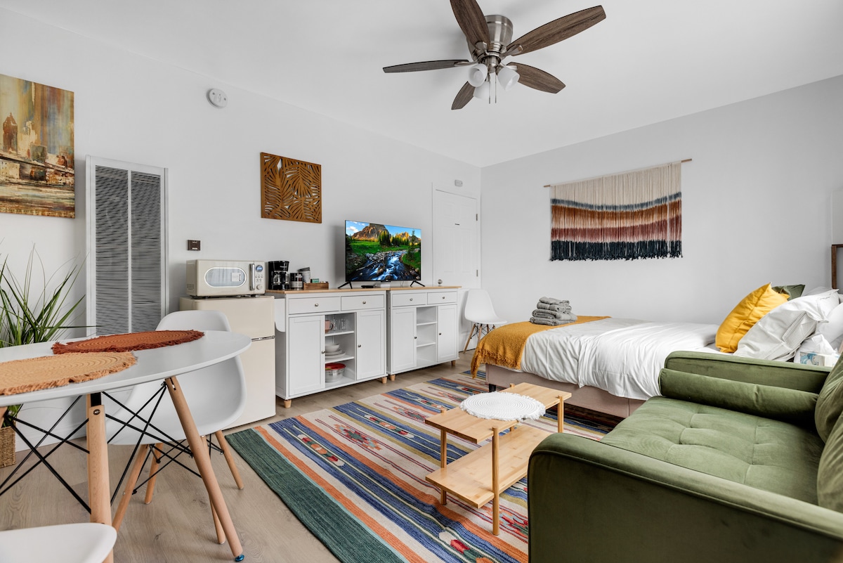 The studio features a queen bed draped with light linens, a comfortable green sofa, and a warm-toned rug. A kitchenette is positioned alongside a dining area with a round table. A large television is mounted on a white unit, and natural light illuminates the space.