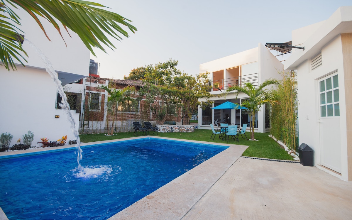 A clear view of a refreshing swimming pool surrounded by tropical landscaping. The pool features a fountain, and there are sun loungers and an umbrella nearby. A white house with multiple levels is visible in the background, complemented by lush greenery.