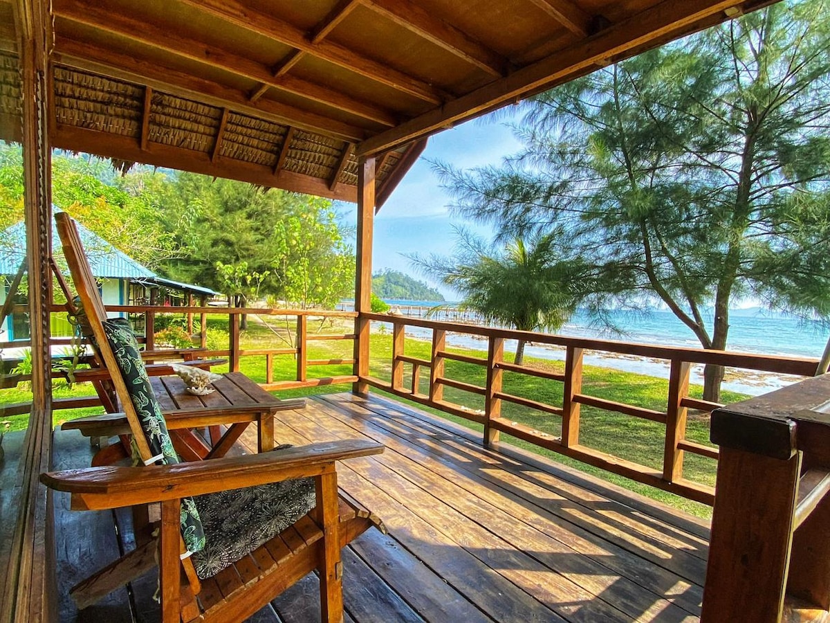 A wooden deck features comfortable seating arrangements, providing views of the shoreline and surrounding greenery. The rustic structure is shaded by a thatched roof, inviting relaxation while enjoying the sights and sounds of nature.