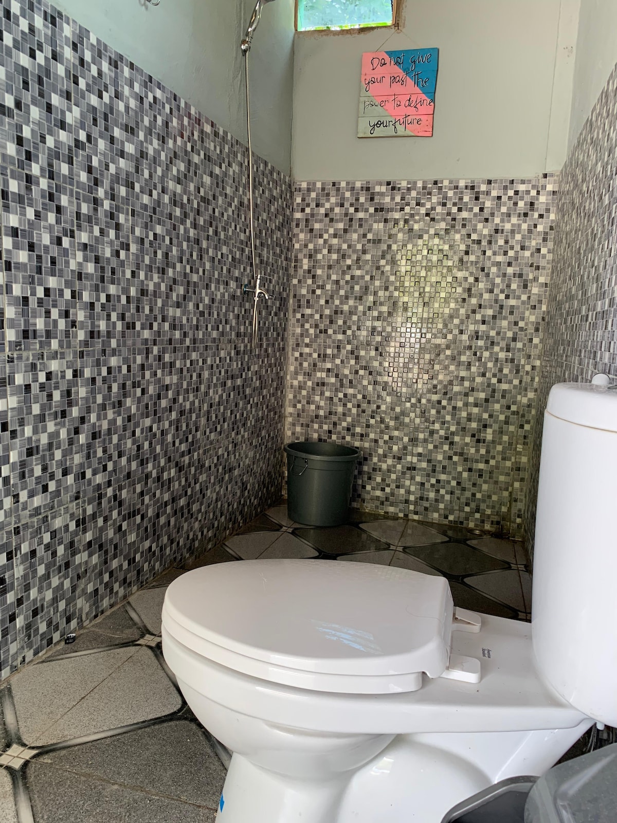 A bathroom is presented with textured mosaic tiles on the walls and patterned flooring. A white toilet is positioned in the lower right corner, while a small green waste bin can be seen nearby. A window is located above, allowing natural light to enter the space.
