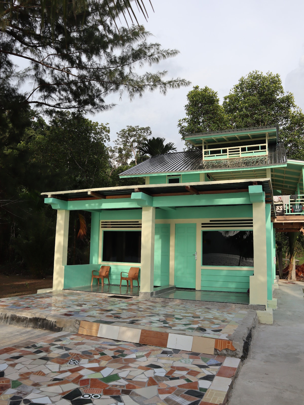 The exterior of a two-story building features a vibrant light green facade and large windows. A small balcony is visible on the upper level, while two wooden chairs are positioned on a mosaic-tiled patio in front of the house.
