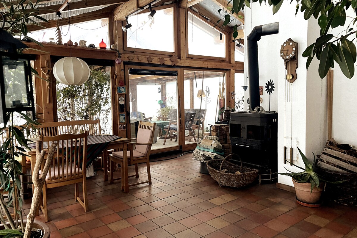 A bright winter garden is featured, showcasing large glass doors that invite natural light. Wooden furniture, including a table and chairs, complements the terracotta-tiled floor. A wood stove and various plants add a cozy touch to the space.