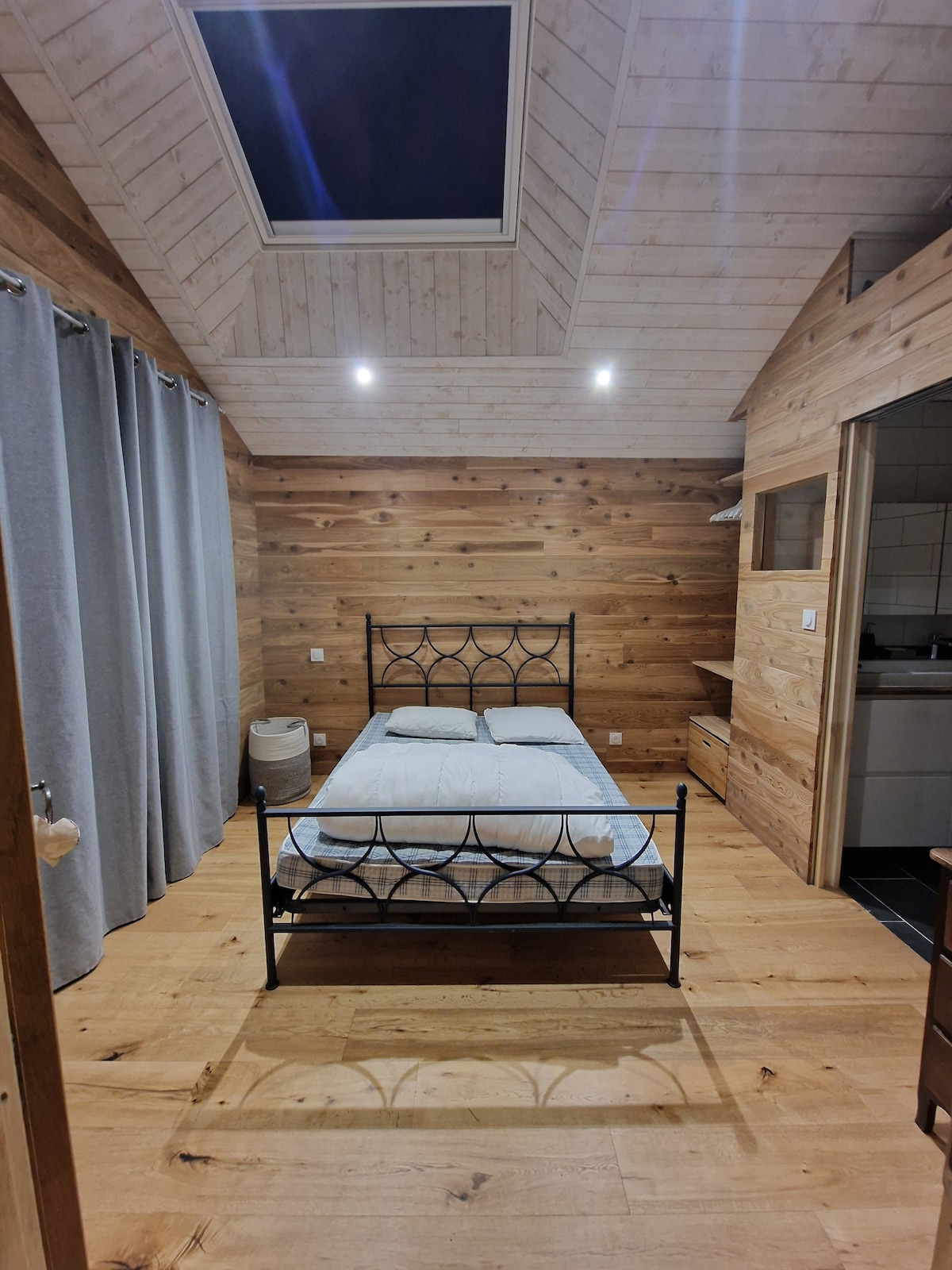 A minimalist bedroom is showcased with wooden walls and a light wooden floor, emphasizing a rustic charm. A spacious bed with a dark metal frame is central, and natural light is provided by a skylight. Soft gray curtains frame the window, contributing to the calming atmosphere.