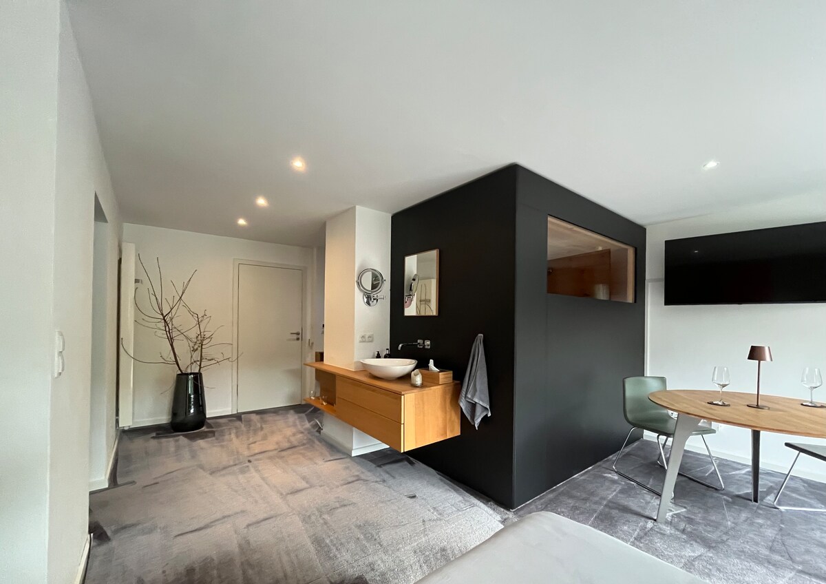 An open living area features a minimalist design with a dark wall section showcasing a mounted mirror. A wooden vanity with a basin is positioned beside a door. A round dining table is accompanied by modern chairs, while a wall-mounted TV is visible to the side.