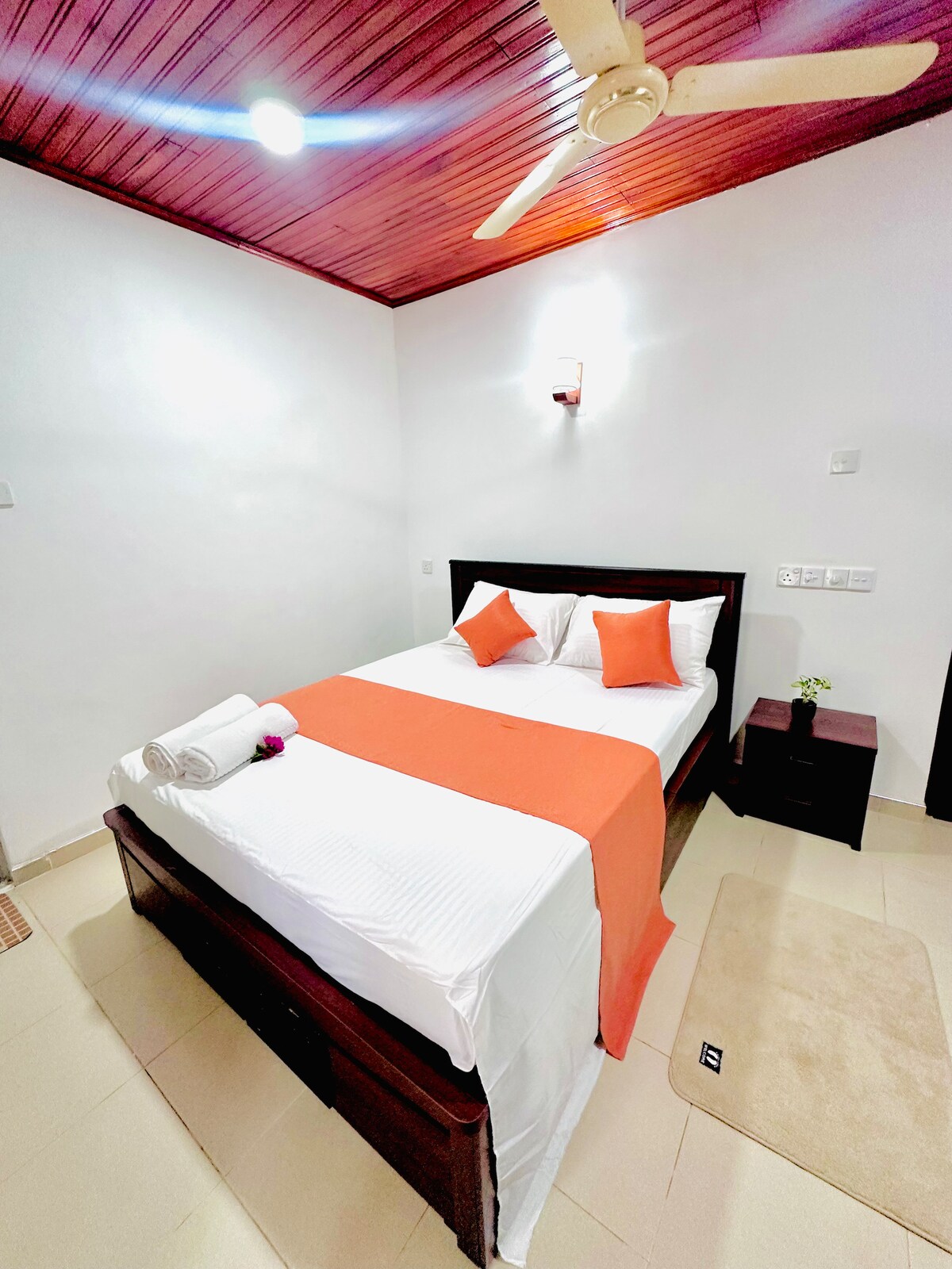 A simple sleeping area features a spacious bed dressed in white linens, accented by two orange pillows. A small nightstand holds a decorative flower, and a light fixture illuminates the white walls. A ceiling fan provides ventilation in the well-lit room.