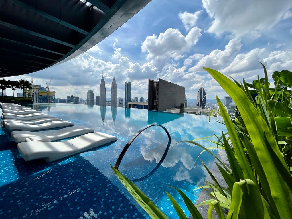 A rooftop infinity pool reflects the surrounding skyline under a partly cloudy sky. Lounge chairs are positioned nearby, and lush green foliage adds a touch of nature. The cityscape includes tall buildings, creating a vibrant urban atmosphere.