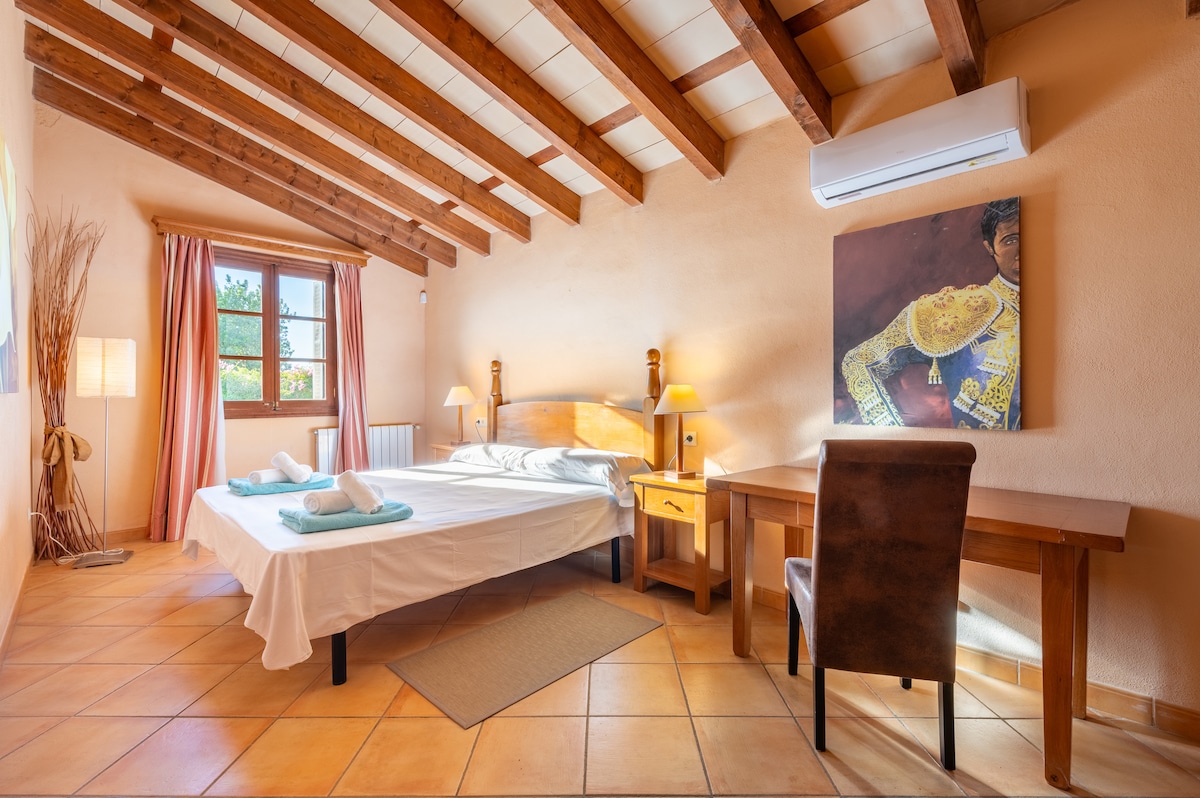 A spacious bedroom is featured, showcasing a large bed with light bedding. Natural light enters through a window framed by soft curtains. Wooden beams accentuate the ceiling, while a desk and a chair are positioned nearby, offering a functional workspace.