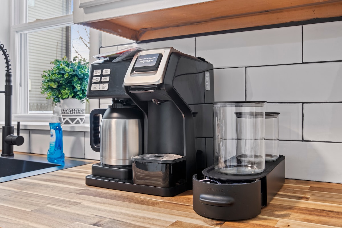 A coffee station is featured on a wooden countertop. A modern coffee maker is positioned beside a stainless-steel carafe, with clear containers for coffee grounds located nearby. Natural light filters in through a window, highlighting a small green plant on the windowsill.