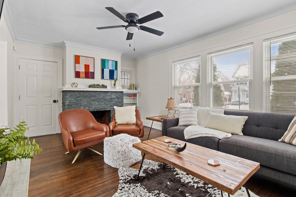 A bright living room features a dark gray sofa complemented by two brown leather armchairs. A wooden coffee table sits on a textured rug, while a stone fireplace is adorned with colorful wall art. Natural light filters through multiple large windows, enhancing the inviting space.