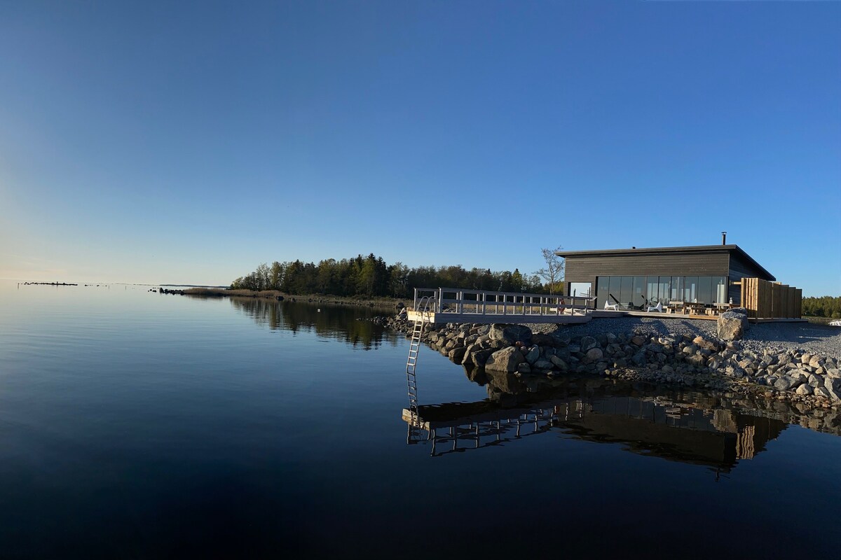 A modern structure sits on the edge of a tranquil body of water, reflecting the clear sky above. Stone steps lead into the water, while a wooden deck extends from the building, providing a serene outdoor space. Lush greenery is visible in the background.