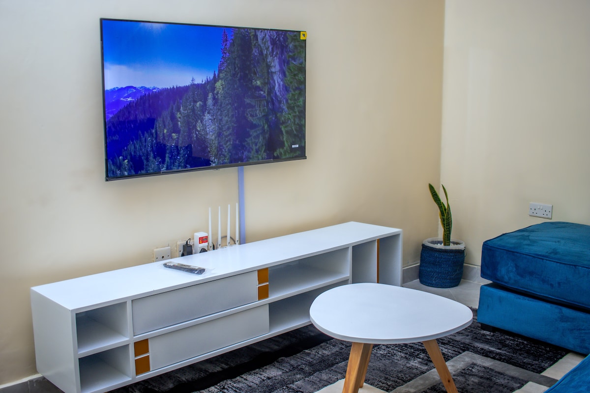 A living area features a modern white TV stand with a large flat-screen television displaying a scenic landscape. A round coffee table with wooden legs is positioned on a patterned rug, alongside a comfortable blue couch. Decorative plants are situated in stylish pots.