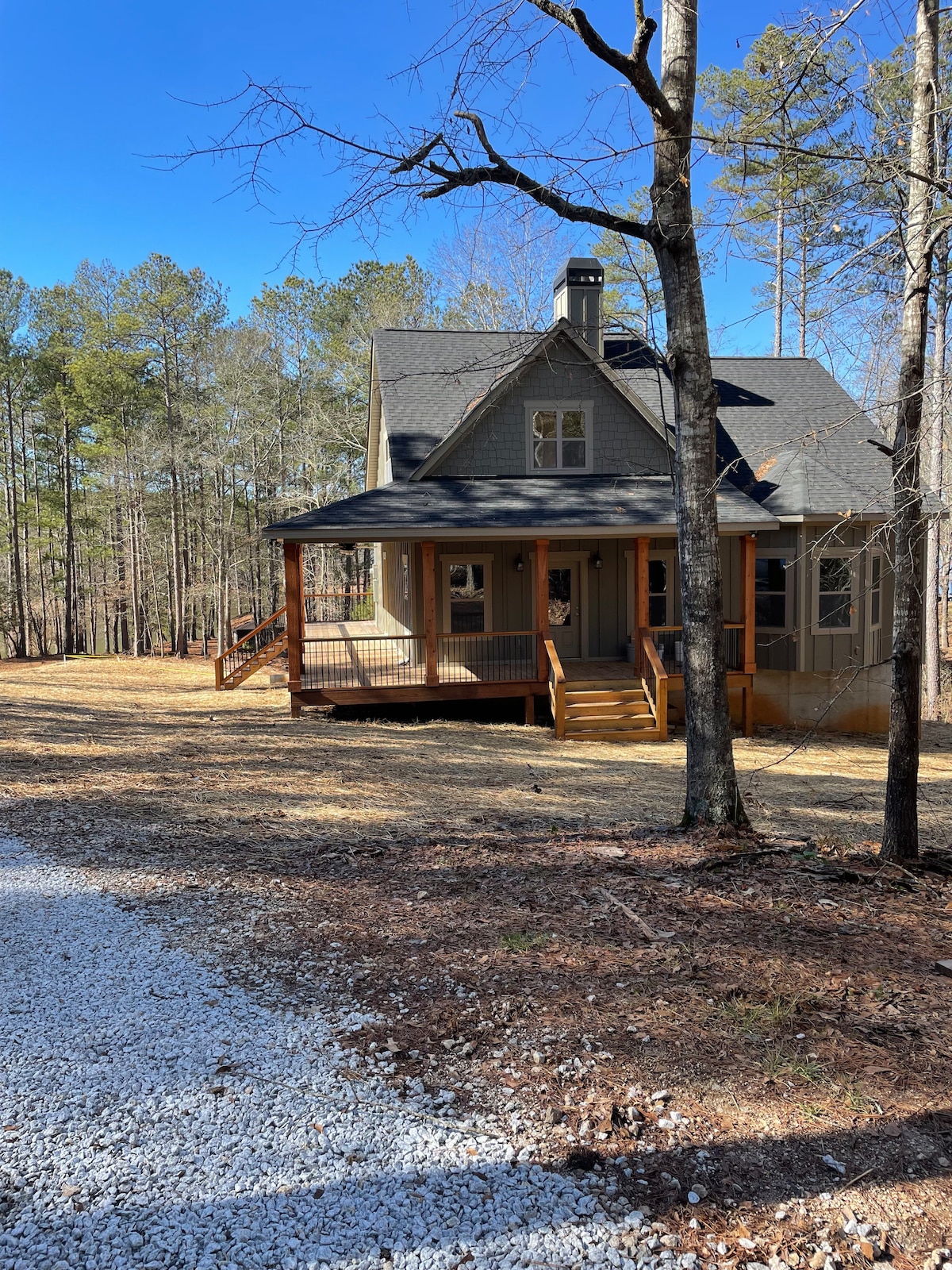 A charming house is situated amidst tall trees, with a welcoming porch and steps leading to the entrance. The exterior features a mix of gray siding and dark shingles, complemented by a lightly gravelled driveway that extends through a wooded surrounding.
