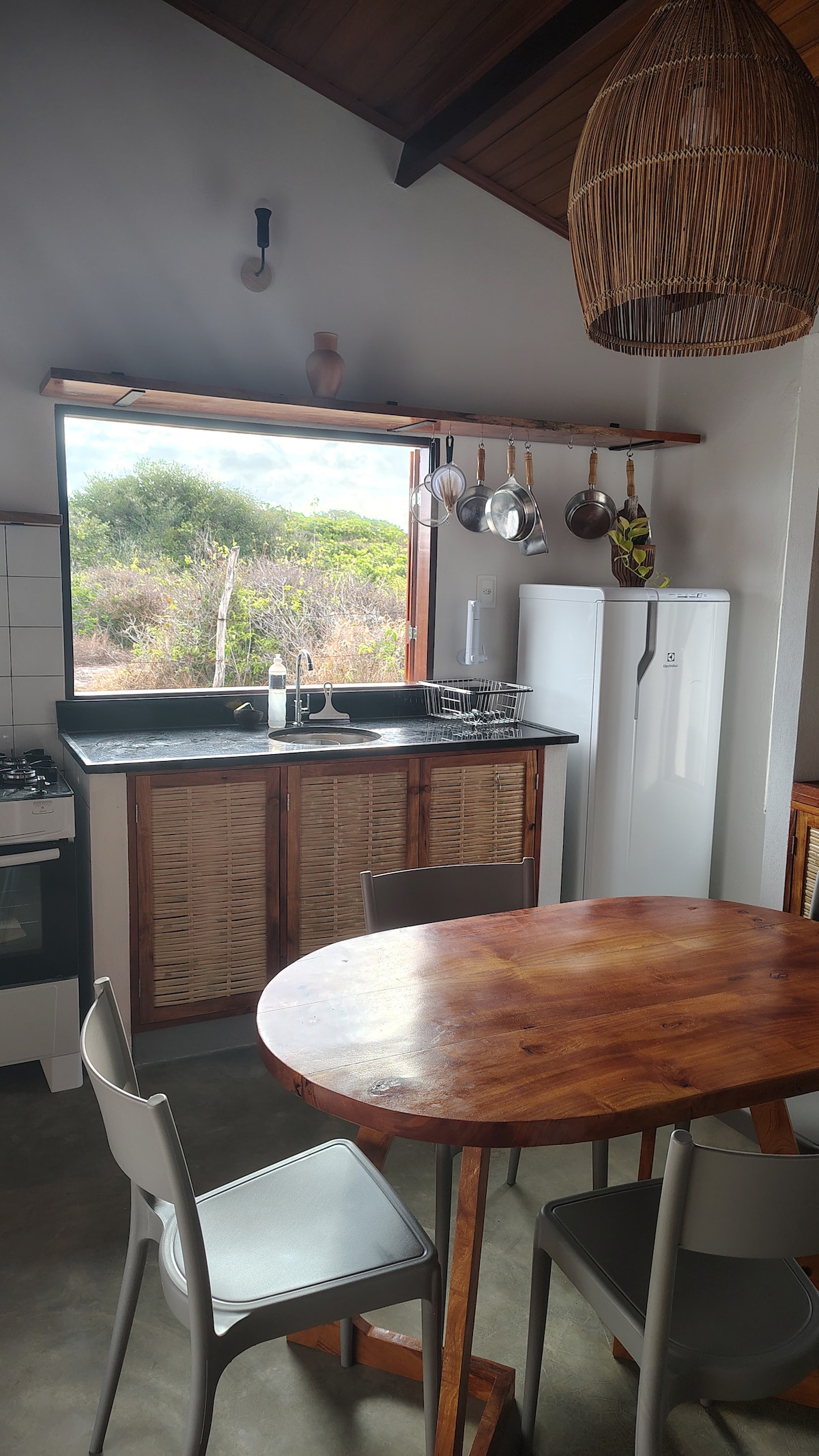 A functional kitchen space features natural light streaming through a large window, offering views of surrounding greenery. Wooden cabinetry with woven accents is complemented by a circular dining table surrounded by chairs. Kitchen appliances, including a stove and refrigerator, are neatly arranged.