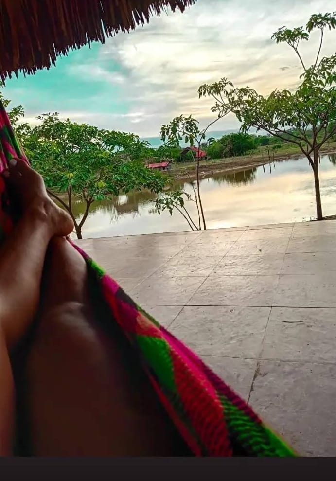 A hammock is depicted, with feet gently resting inside. In the background, lush trees frame a serene body of water, reflecting the soft colors of the sky. The scene suggests a peaceful retreat, ideal for relaxation.