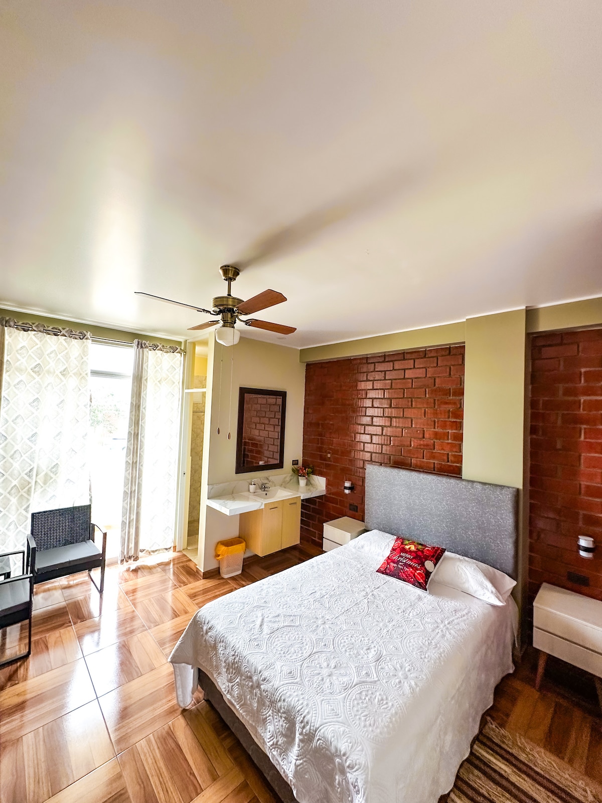 A well-appointed room features a spacious bed with a light-colored coverlet. A seating area includes two chairs and a small table. Natural light enters through large windows, complemented by sheer curtains. The room has a unique exposed brick accent wall and a ceiling fan.