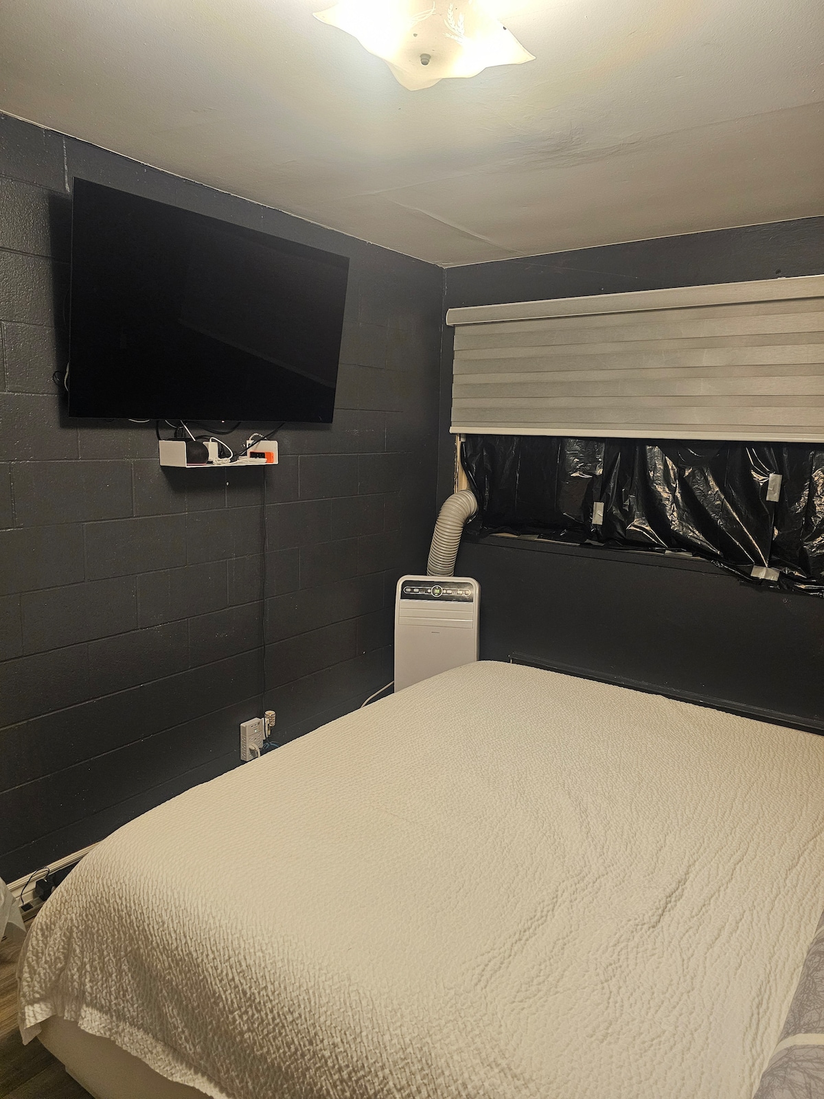A cozy sleeping area is provided, featuring a spacious bed covered with a light-colored blanket. A flat-screen TV is mounted on the wall, above a compact air conditioning unit. The window is covered with shades, allowing for privacy and control over natural light.
