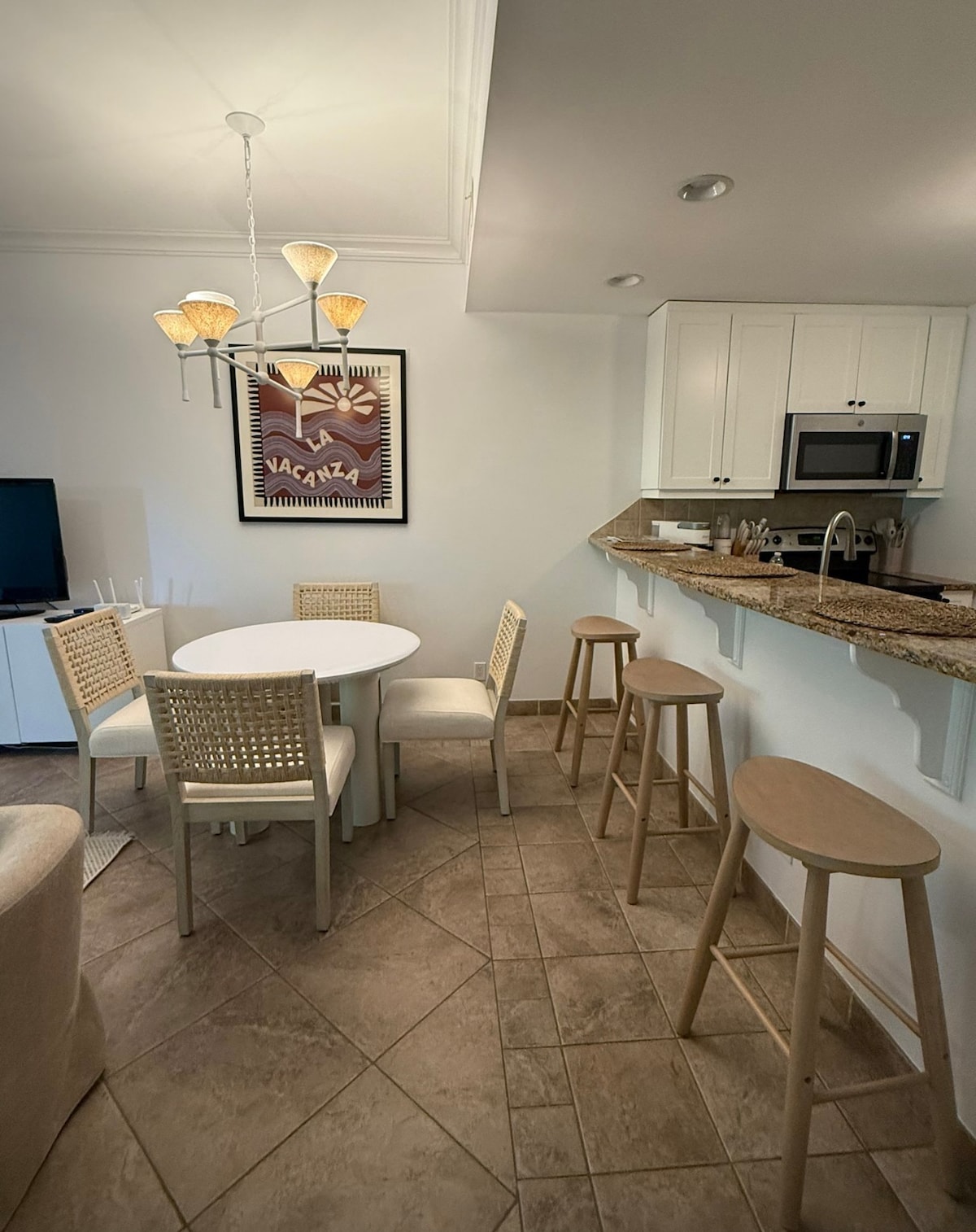 A dining area features a round white table surrounded by four chairs, complemented by two wooden bar stools at the counter. A stylish light fixture hangs overhead, and a small television is positioned in the corner. The kitchen area is visible in the background.
