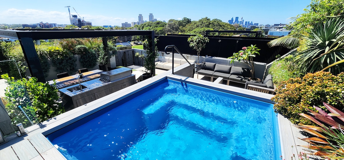 A private rooftop oasis features a sparkling pool surrounded by lush greenery and contemporary outdoor furniture. An outdoor kitchen with BBQ facilities is visible, flanked by a shaded pergola. City skyline views can be seen in the background, enhancing the space's appeal.