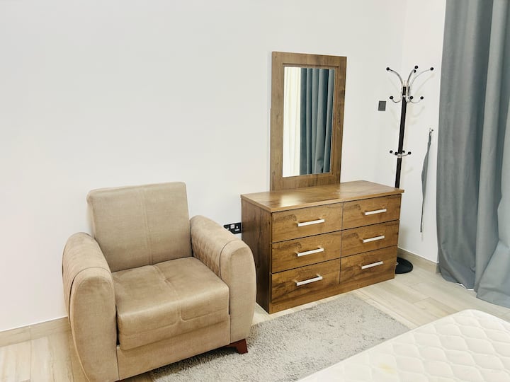 Esquina del dormitorio con un sillón beige colocado junto a una cómoda de madera con seis cajones. Hay un espejo rectangular montado encima de la cómoda. Hay un perchero negro junto a la cómoda, cerca de un conjunto de cortinas de color gris azul. 