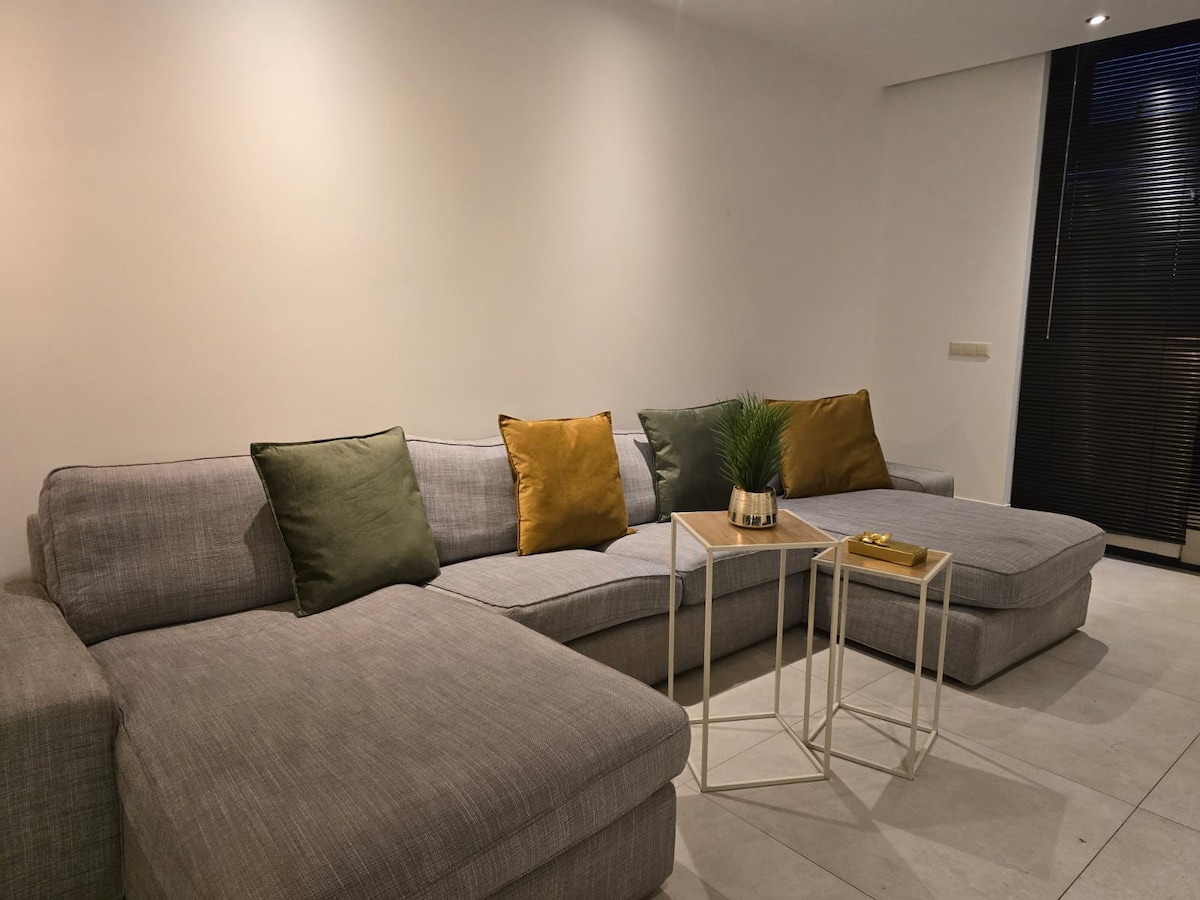 A spacious sectional sofa is featured, adorned with two colored cushions. A minimalist side table is positioned nearby, holding a small plant and a decorative item. Soft lighting enhances the clean lines of the room, complemented by a large window with horizontal blinds.