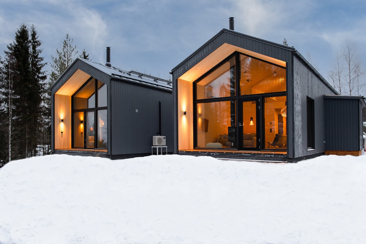 Two modern chalets are nestled in a snowy landscape, featuring large glass windows that reflect warm indoor lighting. The contrasting dark exteriors are highlighted by warm tones from the illuminated interiors, enhancing the inviting appearance against the serene winter backdrop.