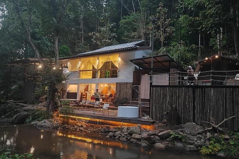 A spacious house nestled among lush greenery features large windows and an inviting outdoor seating area. Warm lights illuminate the structure, creating a welcoming ambiance. A peaceful stream flows nearby, adding to the serene setting. The wooden deck offers a charming spot for relaxation.