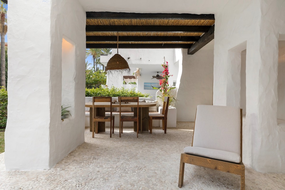 A covered terrace is visible, featuring a wooden dining table surrounded by six chairs. A single wooden lounge chair is positioned nearby. Lush greenery can be seen in the background, adding to the serene setting.