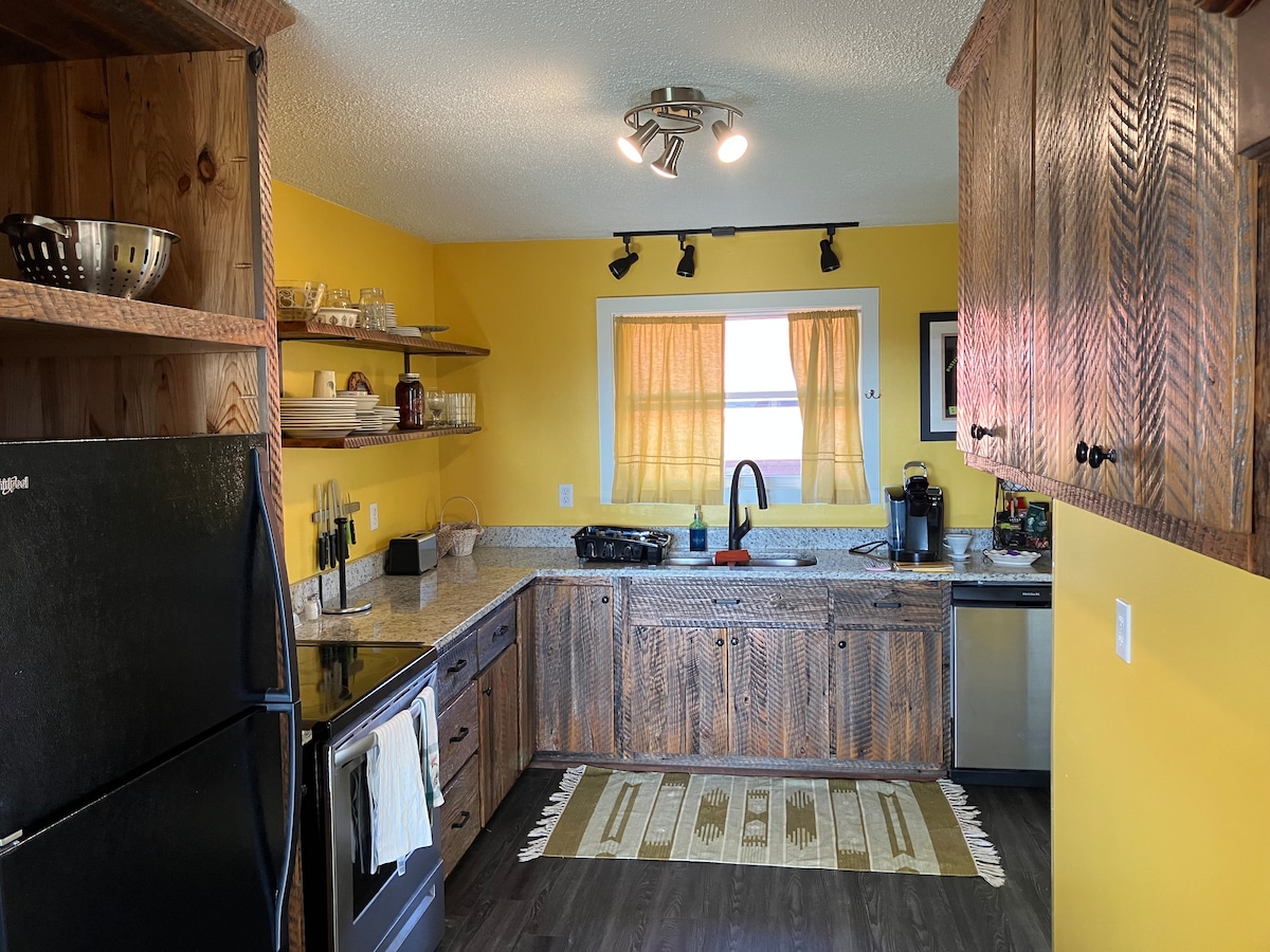 The kitchen features a bright yellow wall, providing a cheerful backdrop. There's a blend of rustic wooden cabinetry and modern appliances. A black refrigerator, stainless steel dishwasher, and a sink with a contrasting red faucet are visible. Natural light enters through a window draped with sheer curtains, illuminating the space.