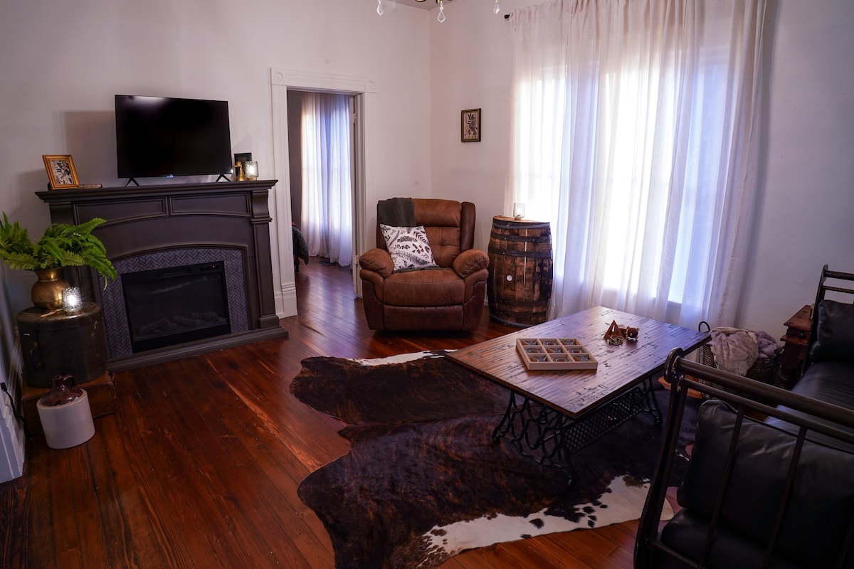 A cozy living area features a plush brown armchair and a dark leather sofa. A wooden coffee table sits atop a patterned rug, and a gas fireplace is framed by decorative elements. Natural light streams through sheer curtains, enhancing the warm ambiance of the space.
