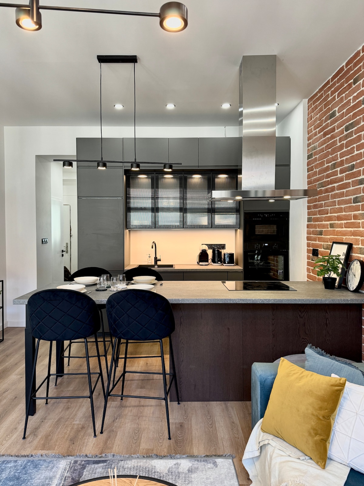 A modern kitchen area features a dark countertop with bar seating for two. Open shelves display various kitchen essentials. A coffee maker and toaster are visible, along with a stylish light fixture overhead and exposed brick accenting the walls, creating an inviting cooking space.