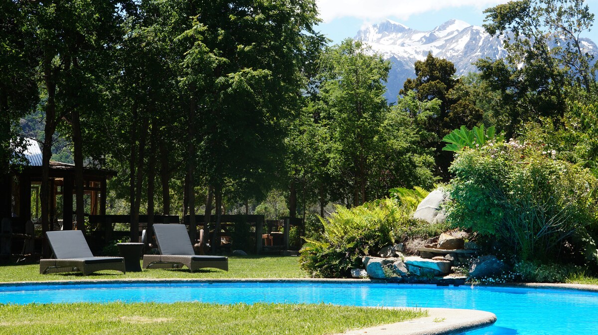 A serene outdoor space features a sparkling blue pool surrounded by lush greenery and tall trees. Comfortable lounge chairs are positioned nearby, inviting relaxation. In the background, majestic snow-capped mountains are visible, enhancing the natural beauty of the setting.