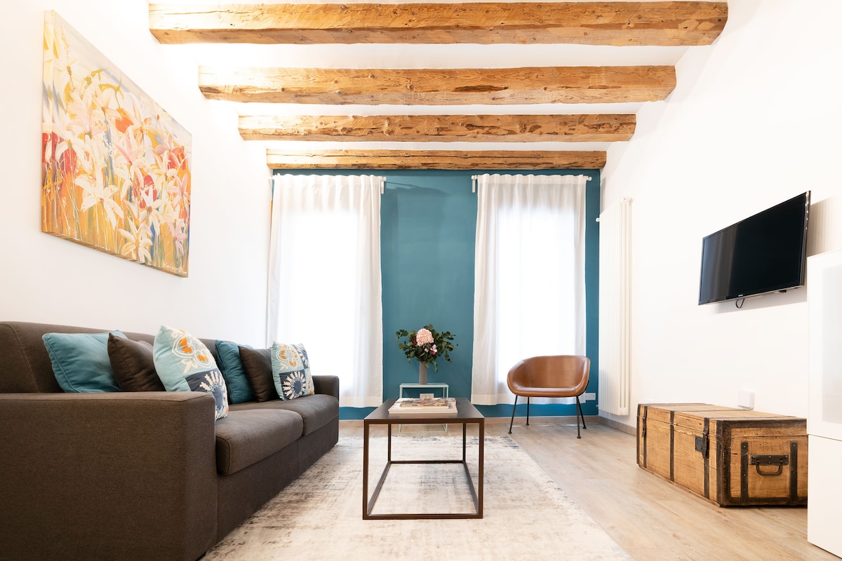 A spacious living area is highlighted by a large artwork above a comfortable dark sofa adorned with colorful cushions. A wooden coffee table sits in the center, and natural light floods in through sheer drapes by the windows, enhancing the room's inviting ambiance.