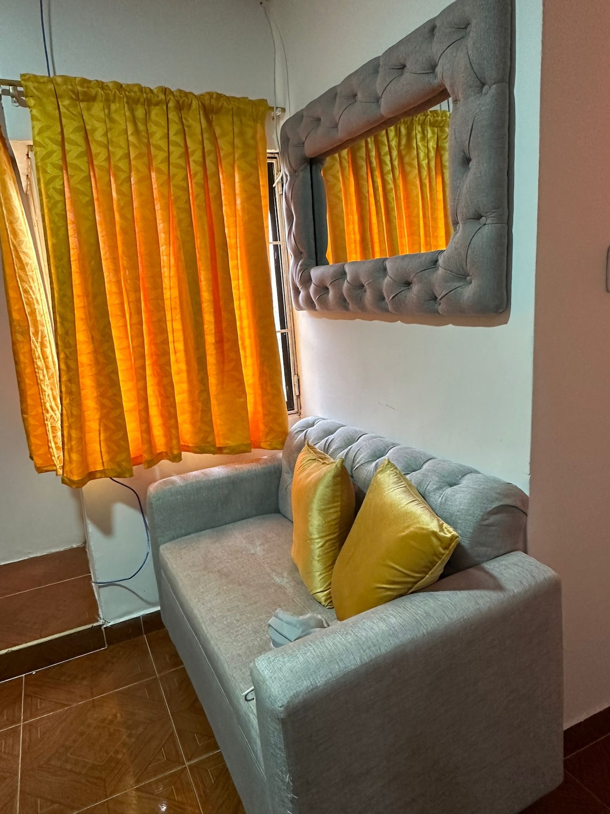 A comfortable gray sofa is positioned against the wall, enhanced by two golden cushions. Natural light filters through a pair of textured yellow curtains, creating a warm atmosphere. A decorative mirror is mounted above the sofa, adding depth to the cozy space.