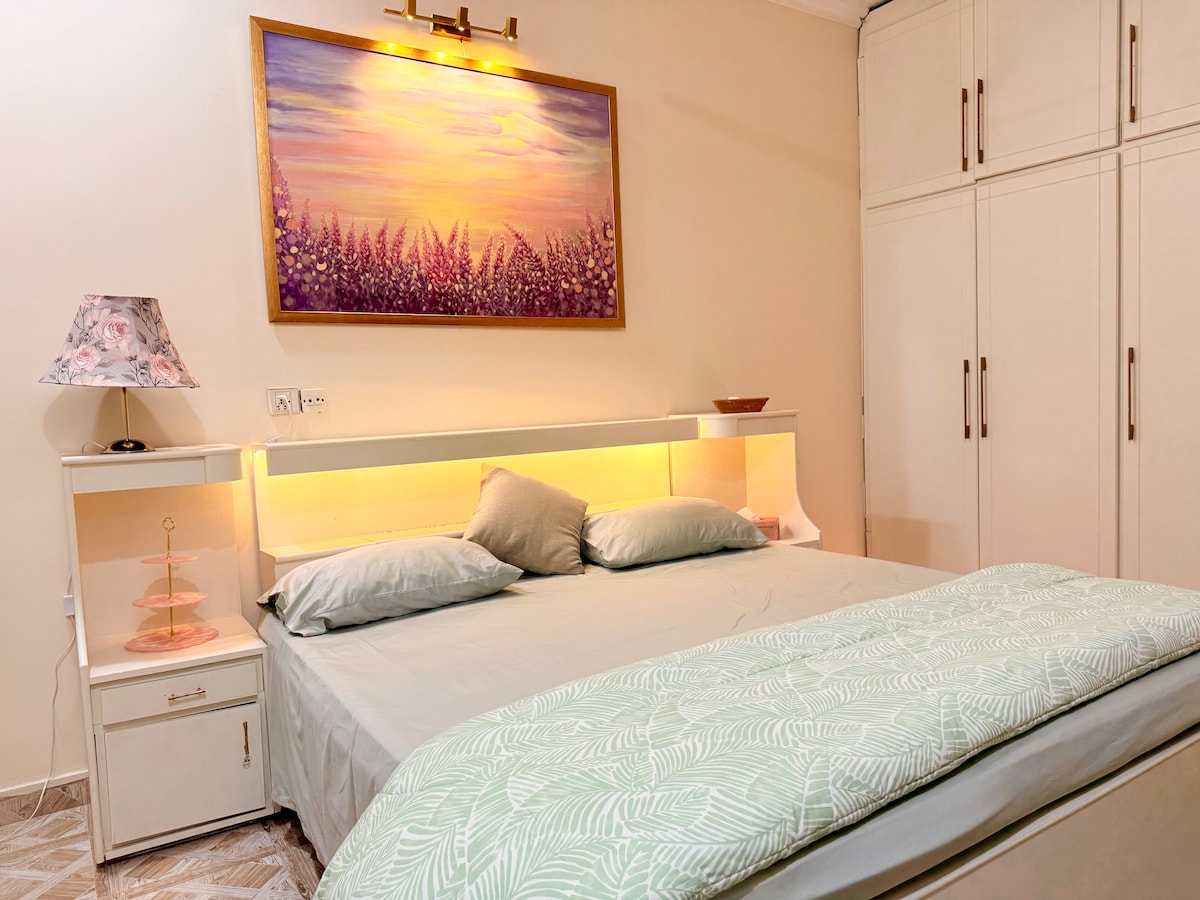 A spacious king-size bed is arranged with soft linens, complemented by decorative pillows. A serene painting depicting a sunset hangs above the headboard. Adjacent cabinetry provides ample storage, while a bedside lamp emits warm light, creating a cozy atmosphere.