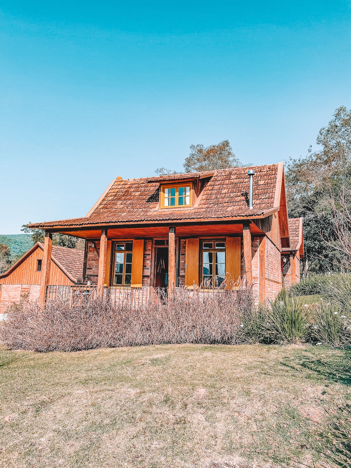 The chalet features a charming facade with warm wooden accents and a sloped roof. The front porch is bordered by lush greenery and bushes, creating a welcoming entrance. Large windows allow natural light to fill the interior while providing views of the surrounding landscape.